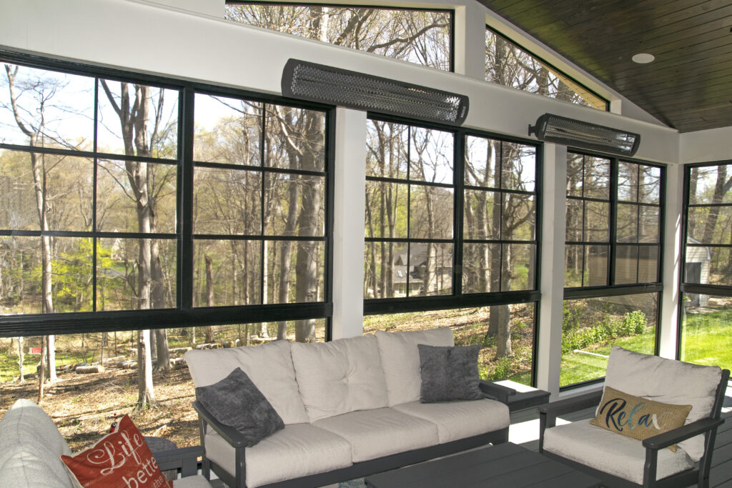 A cozy sunroom with large glass windows featuring a view of a wooded area. The room includes comfortable beige sofas with gray and red accent pillows, a dark coffee table, and ceiling heaters. The atmosphere is serene and inviting.