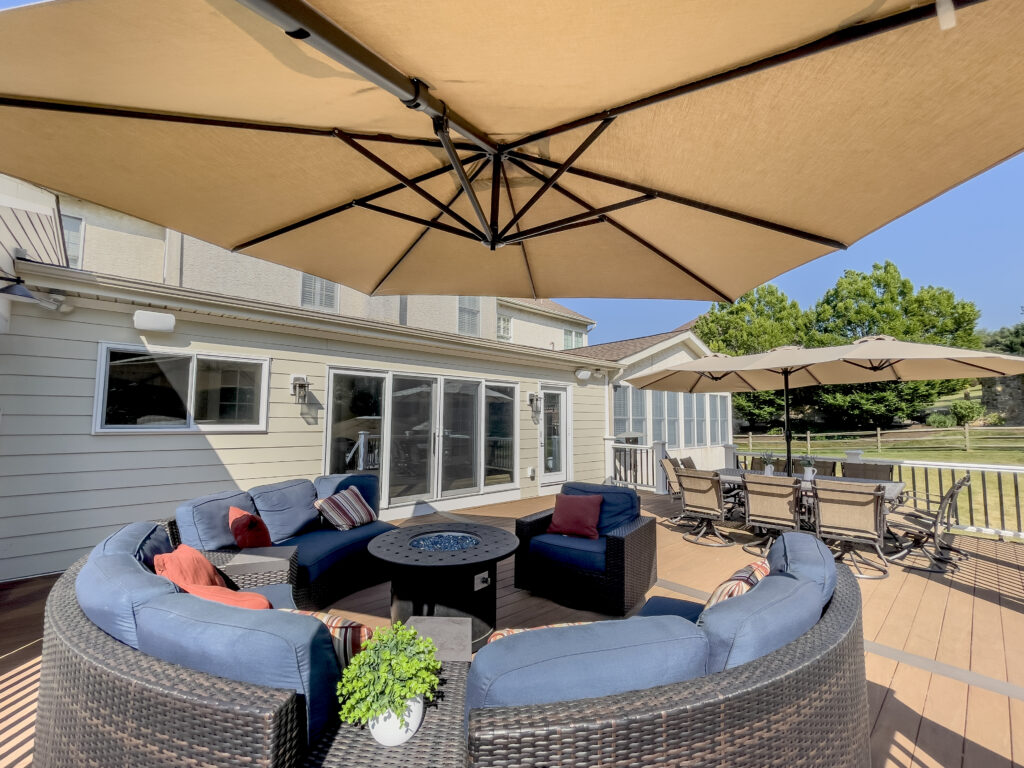 A spacious outdoor patio features a round wicker sofa with blue cushions surrounding a fire pit table. Large beige umbrellas provide shade. A dining table with chairs is in the background, set on a wooden deck adjacent to a house with large windows.