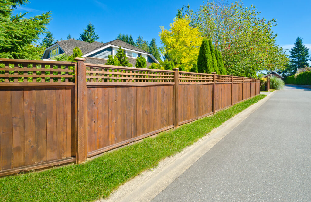 A suburban street with a wooden fence running alongside a neatly paved road. The fence is bordered by a well-maintained green lawn. Behind the fence, trees and a house with a sloped roof are visible under a clear blue sky.