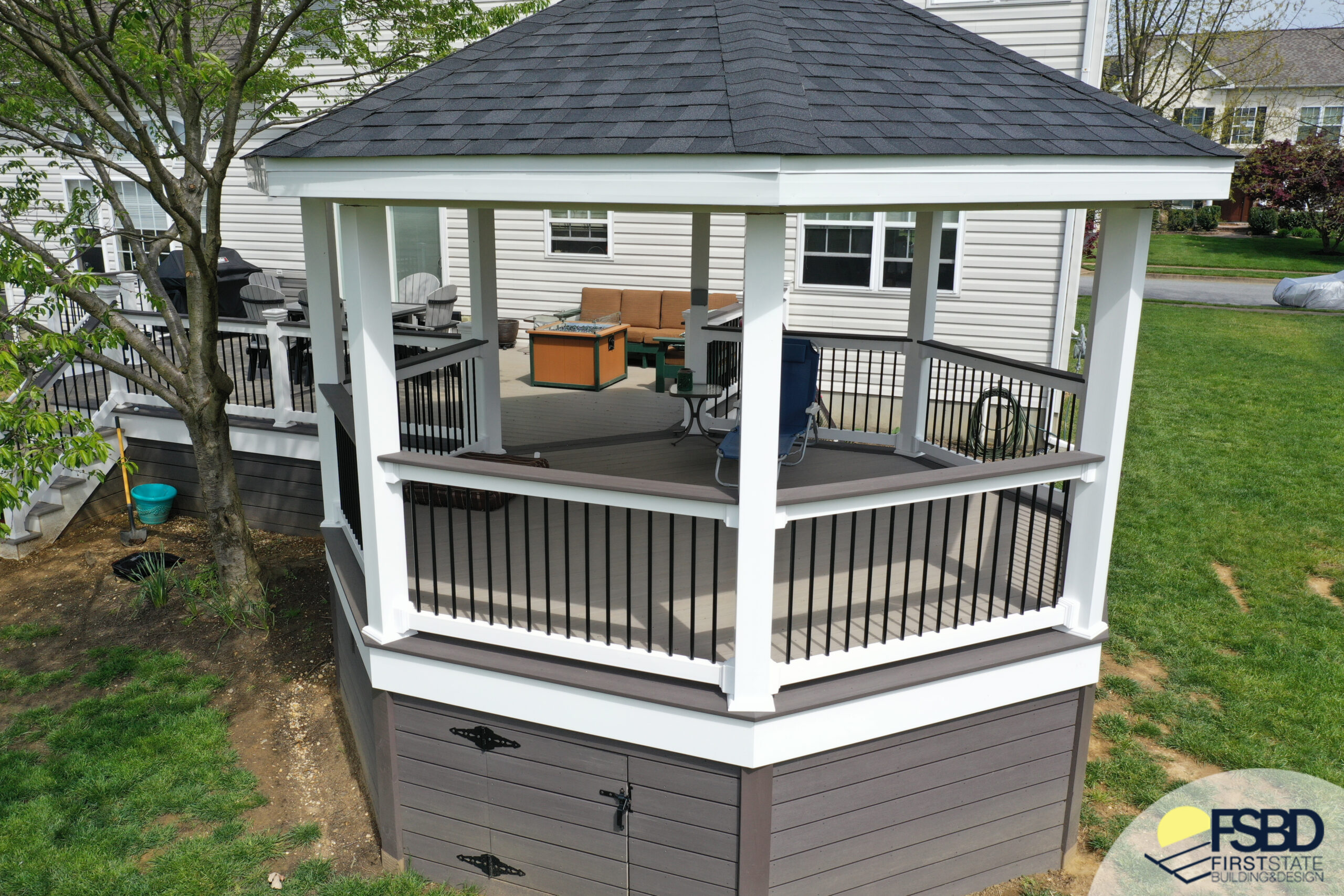 A two-story outdoor gazebo with a black shingled roof and white railing is situated on a lawn next to a house. There are chairs and a small table inside. A First State Building & Design logo is visible in the corner.