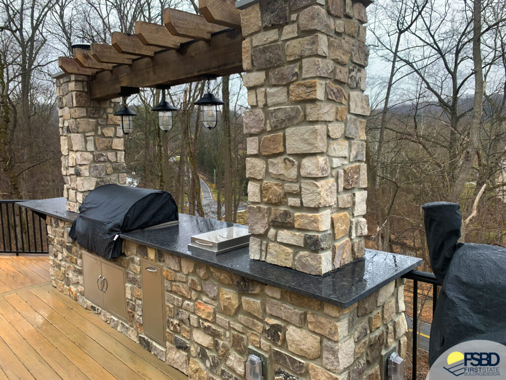 A stone outdoor kitchen with a countertop and grill under a wooden pergola. Three lantern-style lights hang from the pergola. Its a rainy day with trees in the background and a wooden deck in the foreground.