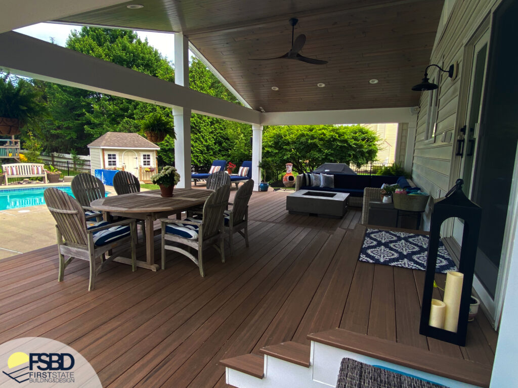 A spacious, covered patio with a wooden floor features a circular dining table with chairs, a cushioned seating area, and outdoor decor. The setting overlooks a lush backyard with a pool and playhouse.