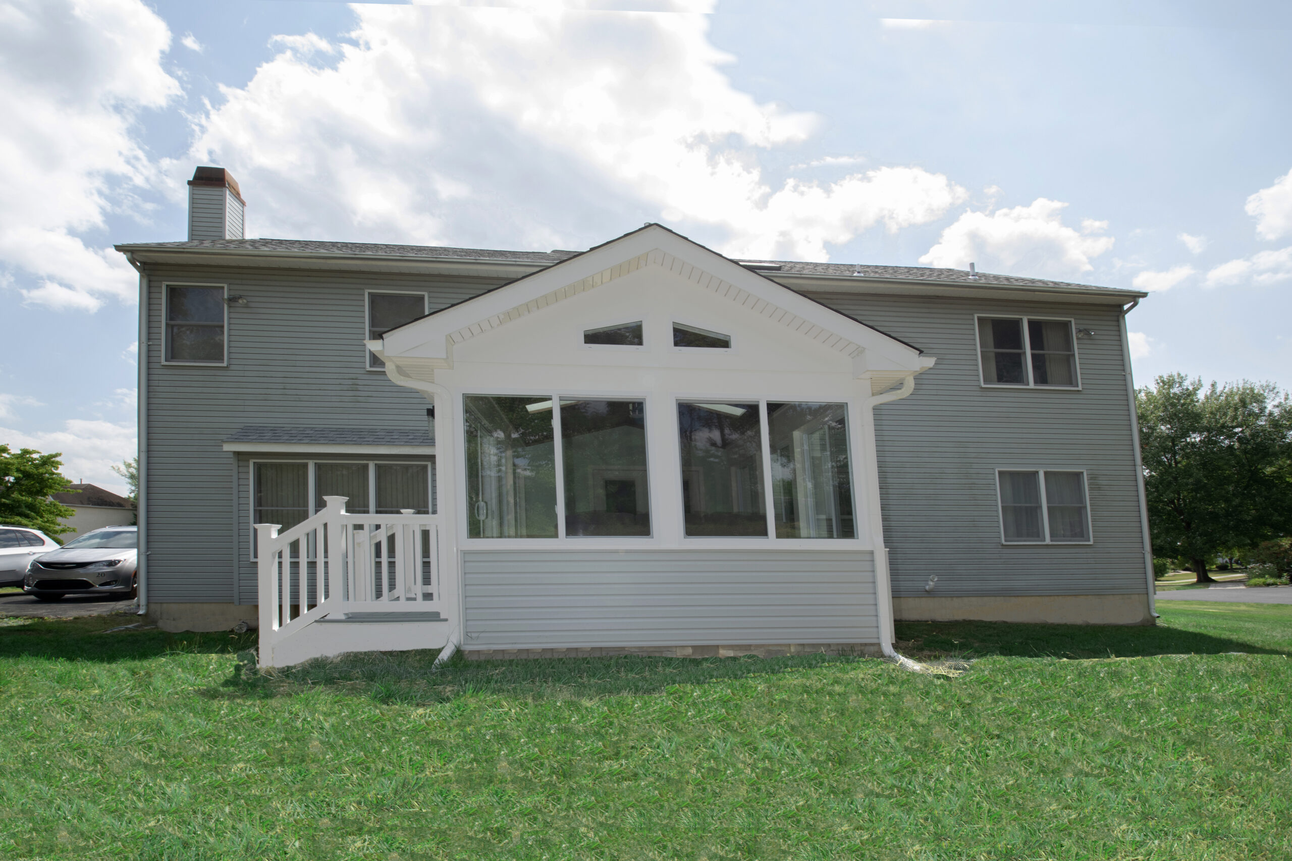 A two-story gray house with a white sunroom extension on a grassy lawn. The sunroom features large windows and a white railing. Partly cloudy sky in the background. A parked car is visible on the left side.