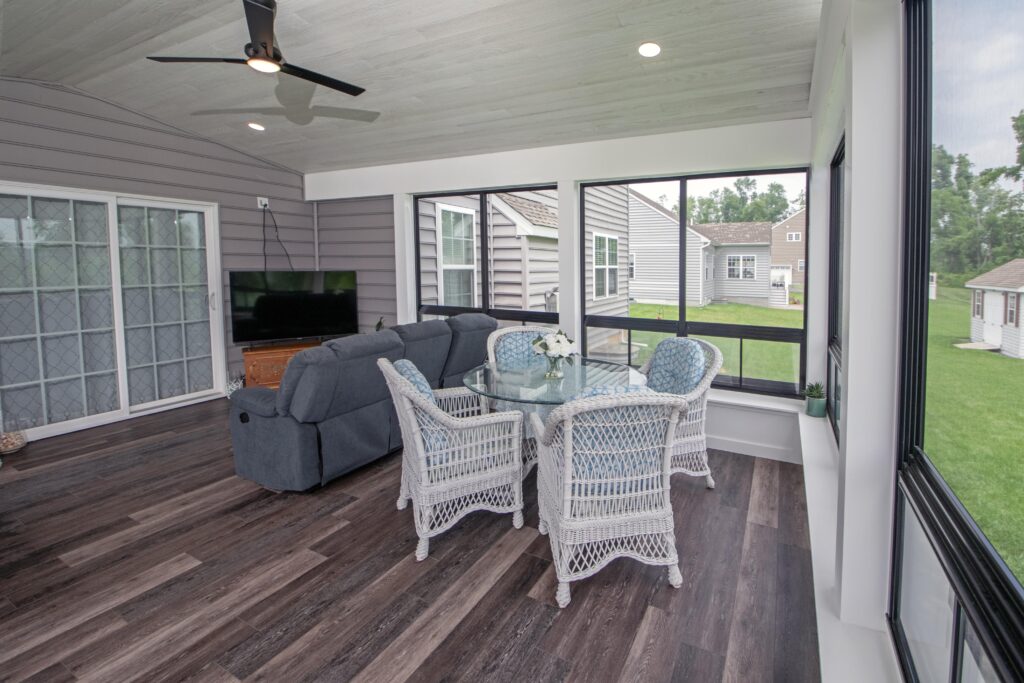 A sunroom with dark wood flooring features a gray couch, a flat-screen TV, and a round glass table with four white wicker chairs. Large windows offer a view of a green lawn and an outdoor building. A ceiling fan is mounted on the white ceiling.