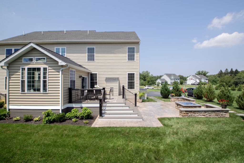 A beige two-story house with a small porch and steps leading to a patio. The backyard features a lawn, potted plants, and a stone patio area. Nearby are trees and a neighboring house under a clear blue sky.