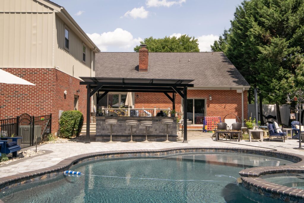 A backyard features a stone patio with a curved swimming pool. A brick house has a covered outdoor bar and a few lounge chairs, with trees and a clear sky in the background.