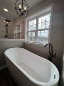 1053 Bathroom 15 | First State Building & Design A modern bathroom featuring a white freestanding bathtub under a window with a view of trees. A black floor-mounted faucet is beside the tub. A glass-enclosed shower with a dark fixture is visible in the background. A black chandelier hangs above.