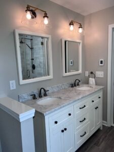 1053 Bathroom 3 | First State Building & Design A modern bathroom featuring a white double vanity with two sinks, marble countertop, and matching mirrors. The gray walls complement the wooden floor. Two light fixtures with glass shades illuminate the area. A glass shower is visible in the background.