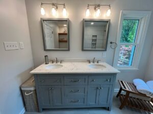 1055 Bathroom 8 | First State Building & Design A modern bathroom features a double sink vanity with a marble countertop and two mirrors. Overhead lighting illuminates the space. A small window on the right offers a view of greenery outside. A wooden bench with rolled towels is nearby.