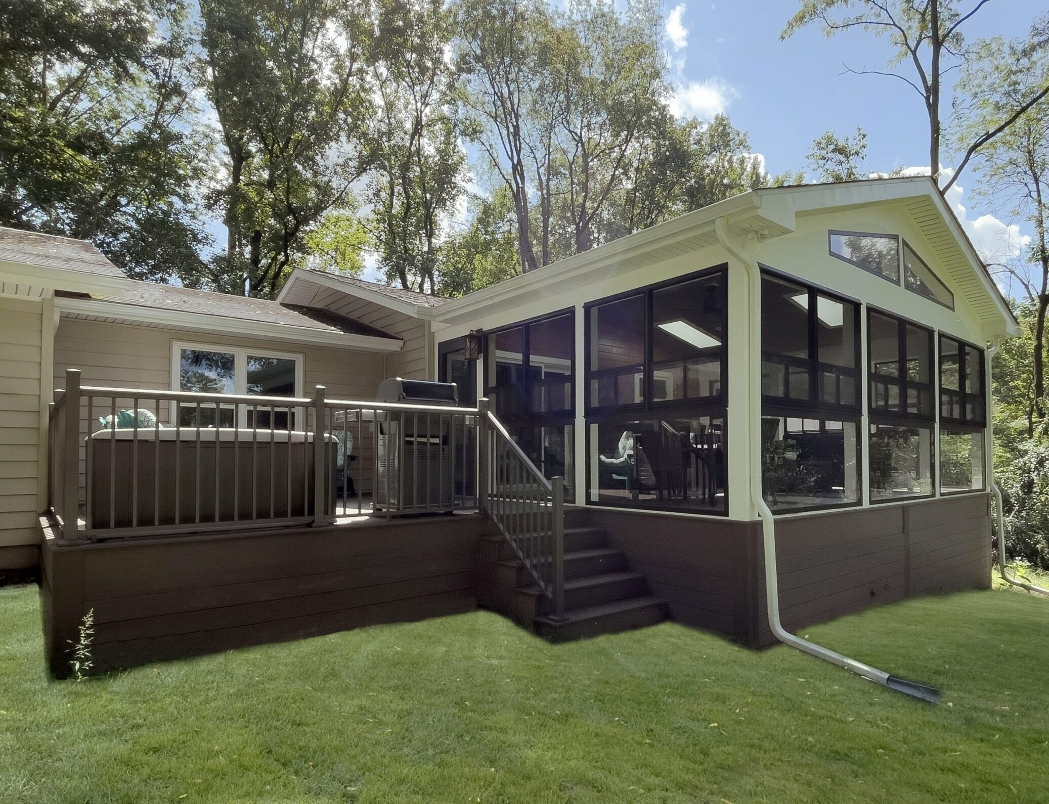 A single-story house with a large sunroom extension featuring dark-framed windows and steps leading to a wooden patio. The house is surrounded by a lush green lawn and tall trees under a partly cloudy sky.