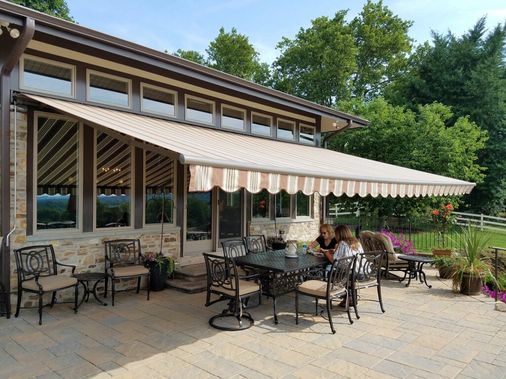 A spacious outdoor patio with a striped retractable awning covering a dining area. Two people sit at a table surrounded by cushioned chairs. The patio is bordered by lush greenery and a stone building with large windows.