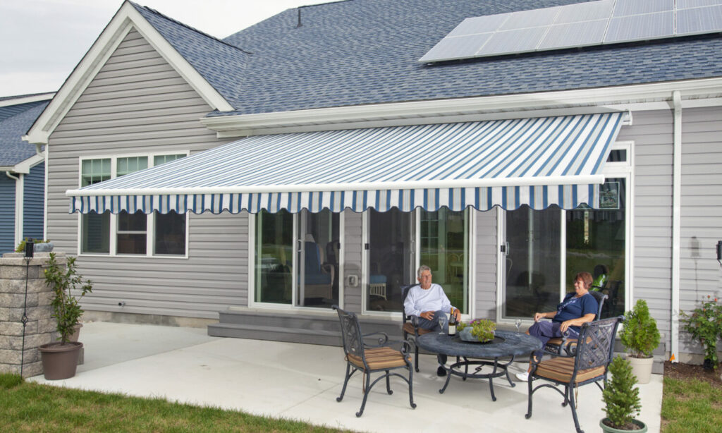 A couple relaxes on a patio under a large, blue-and-white striped retractable awning attached to a gray house. Theres a round table with chairs and small plants around. Solar panels are visible on the roof.