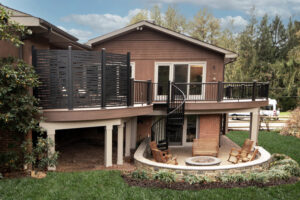 A modern, two-story house with a wooden exterior and a spacious deck featuring a black spiral staircase. Below the deck is a patio area with a fire pit surrounded by wooden chairs, set against a backdrop of trees and grass.