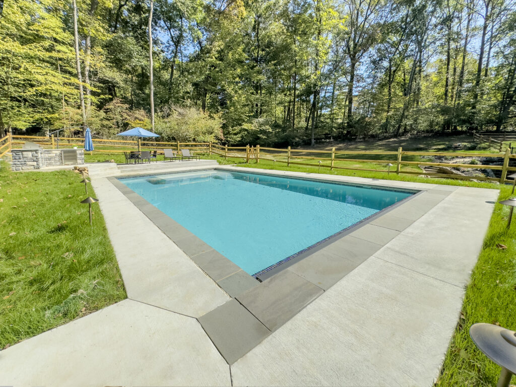 A clear, rectangular swimming pool in a backyard surrounded by greenery and woods. It has a concrete deck with a wooden fence and a blue umbrella over a seating area in the background.