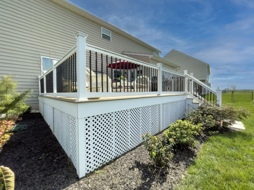 A backyard view featuring a raised wooden deck with white railings and black balusters. The deck has a lattice skirt and is adorned with plants and shrubs. A red umbrella is visible on the deck against a clear blue sky.