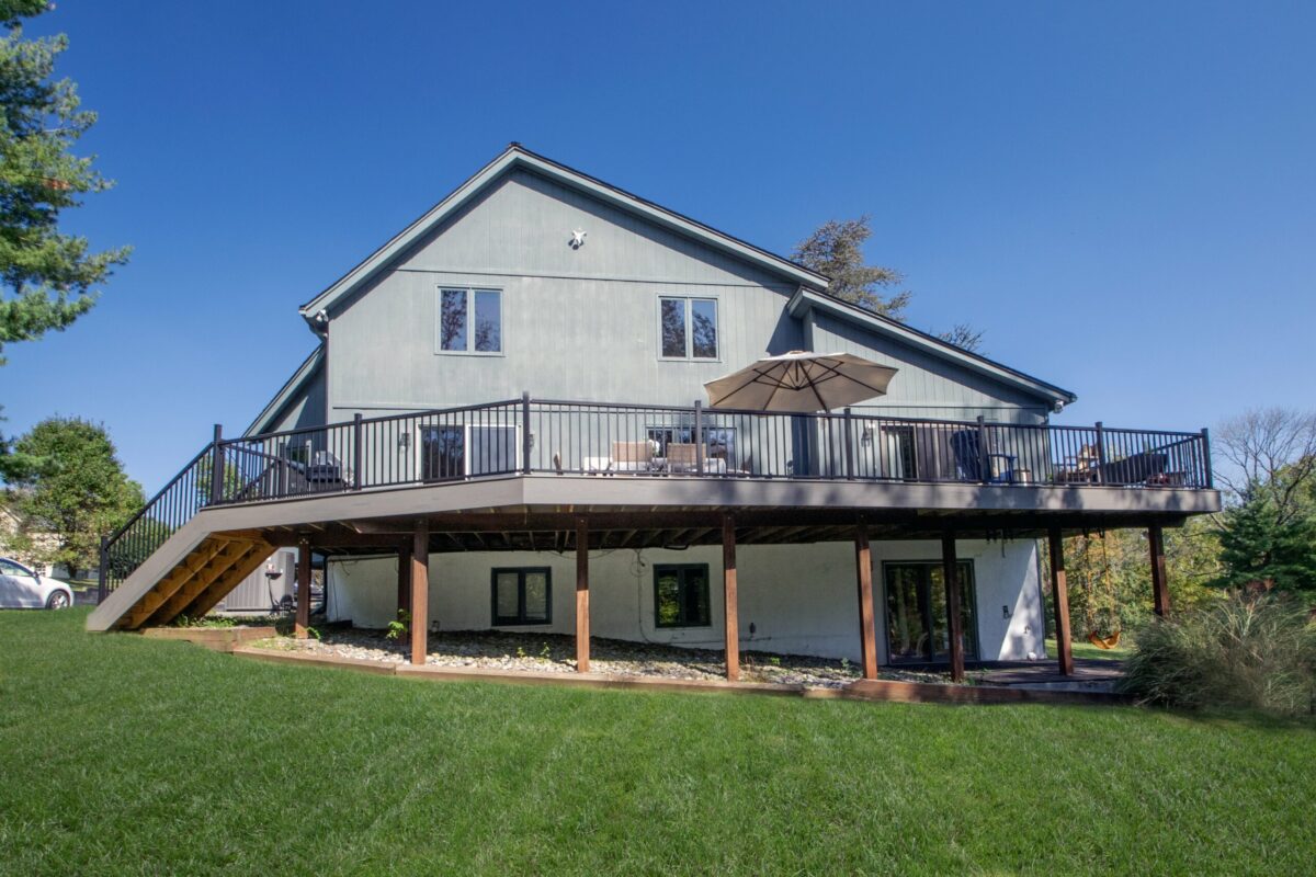 A modern house with a large wooden deck on the upper floor, featuring outdoor furniture and a large umbrella. The deck overlooks a well-maintained lawn and is supported by beams. A staircase leads down to the yard. Clear blue sky in the background.