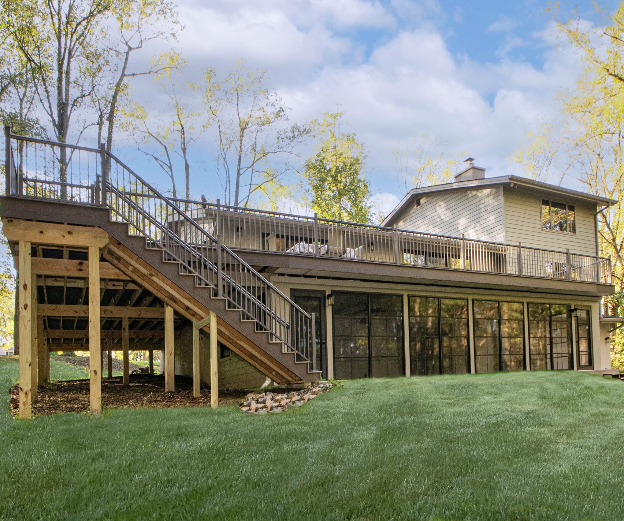 A modern two-story house with a large upper deck and expansive lawn. The house features an under-deck enclosed area with glass panels, situated amidst a backdrop of tall, leafy trees under a partly cloudy sky.