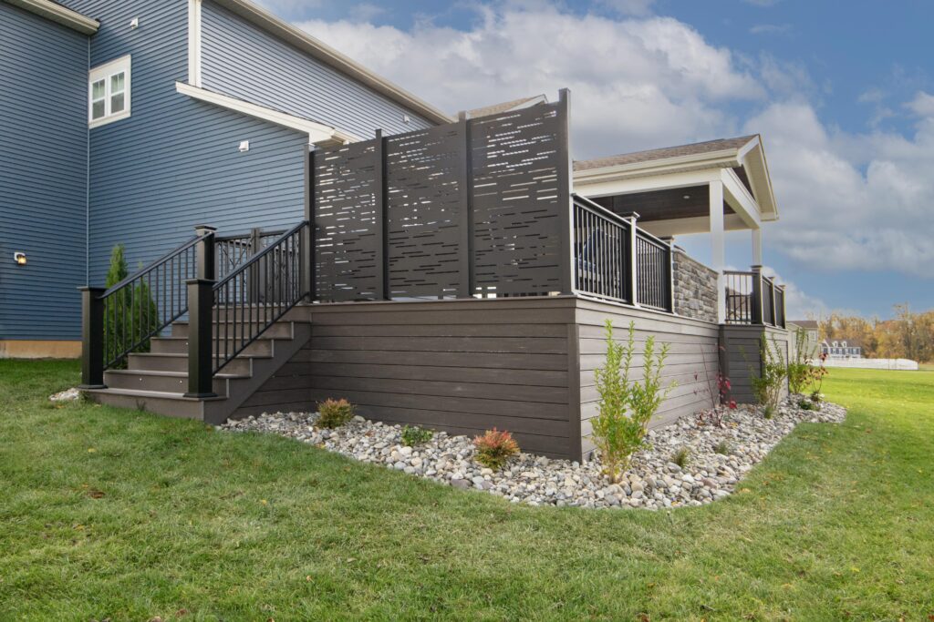 A modern house with a blue exterior features a deck with dark wood railings and a decorative privacy screen. The deck is elevated, with stairs leading up, surrounded by a landscaped area with stones and small plants. The sky is partly cloudy.