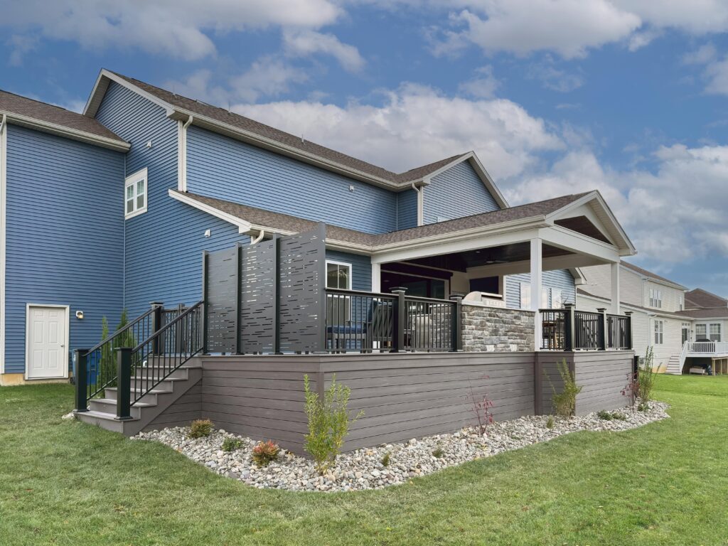 A blue two-story house with an elevated deck featuring outdoor seating, a privacy screen, and a small garden with rocks and plants. The sky is partly cloudy, and the setting is suburban with other houses visible nearby.