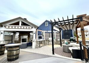 | First State Building & Design Outdoor patio with a pergola, large stone fireplace, and seating area. Includes a wooden bar with barrel bases and contemporary decor. The setting is designed for relaxation and entertainment.