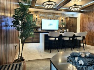 | First State Building & Design A modern living room features a large TV mounted on a stone wall with the words Dream, Design, Build above it. A bar with high-backed chairs faces the TV. The room is warmly lit with decorative ceiling lights and has wooden and stone accents.