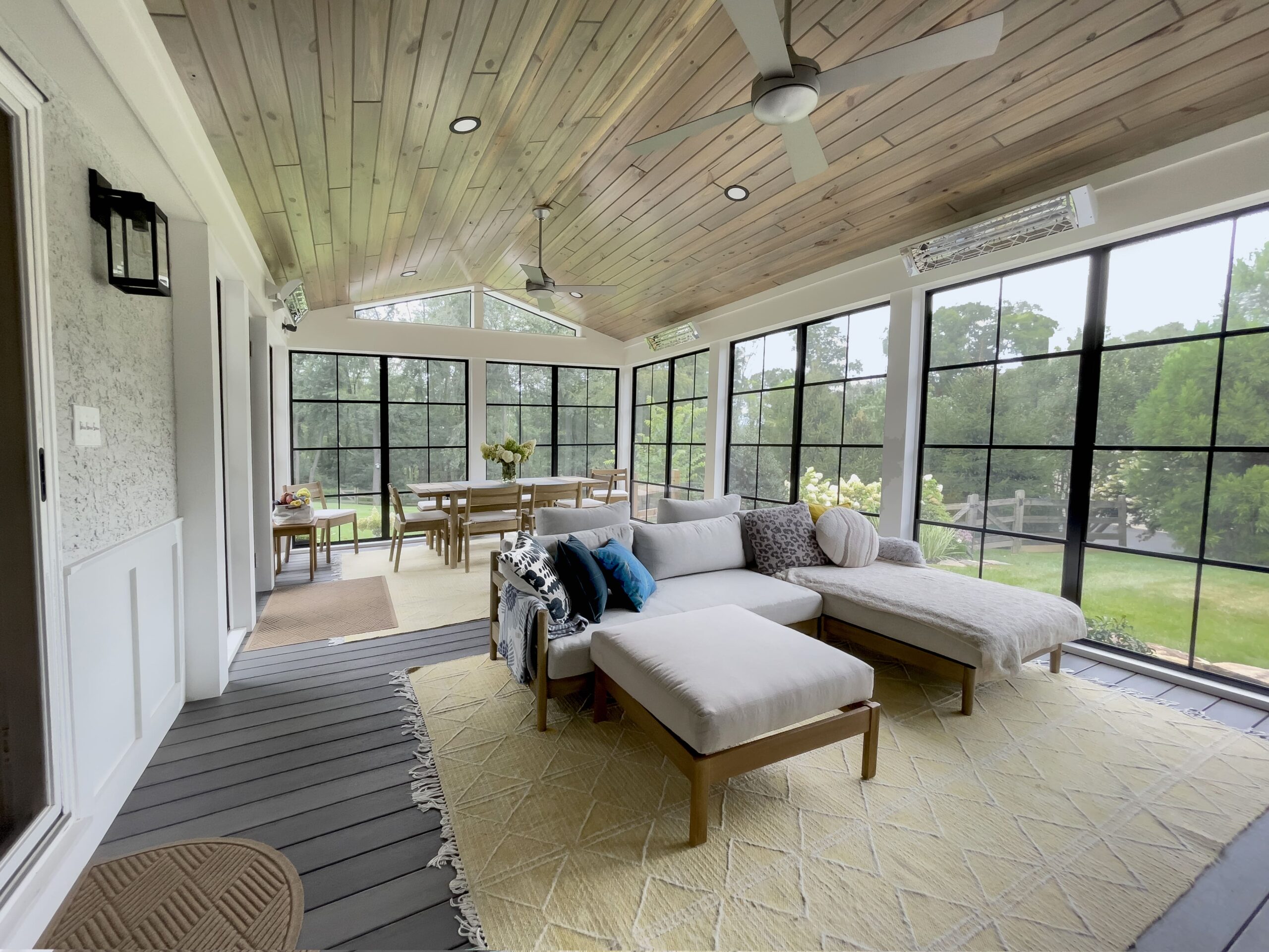 A bright, modern sunroom with a wooden ceiling, large windows, and two ceiling fans. It features a beige sectional sofa on a light geometric rug and a dining table with chairs near the windows, offering a view of lush greenery outside.