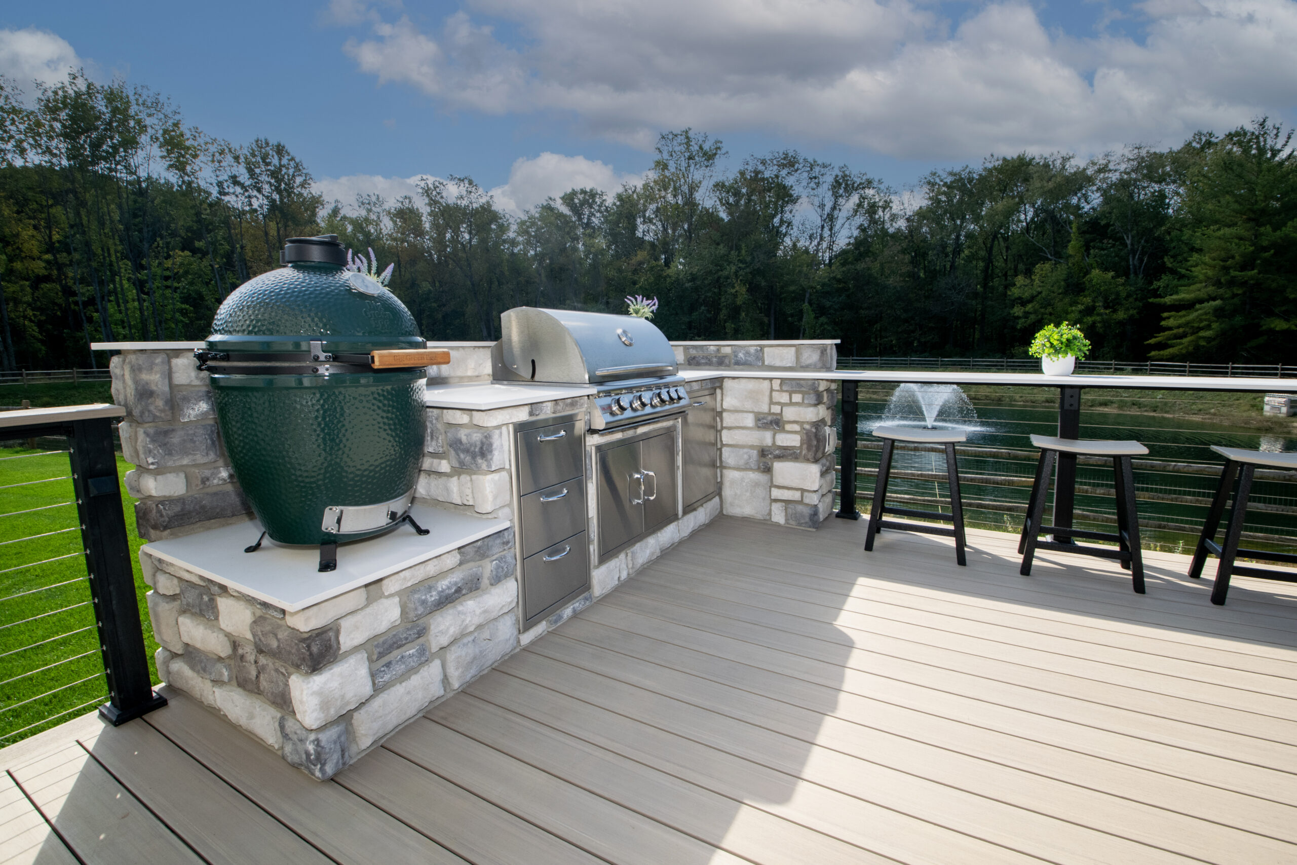 Outdoor deck with a built-in grill station, featuring a green ceramic grill and a stainless steel gas grill. The stone countertops and wooden flooring overlook a grassy area and trees. Two black stools and a small plant decorate the space.