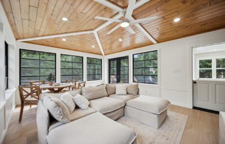 A bright sunroom featuring a comfortable beige sectional sofa and a wooden dining table with chairs. The room has a wooden ceiling, large windows with black frames, and a ceiling fan. Light wood floors and greenery are visible outside.