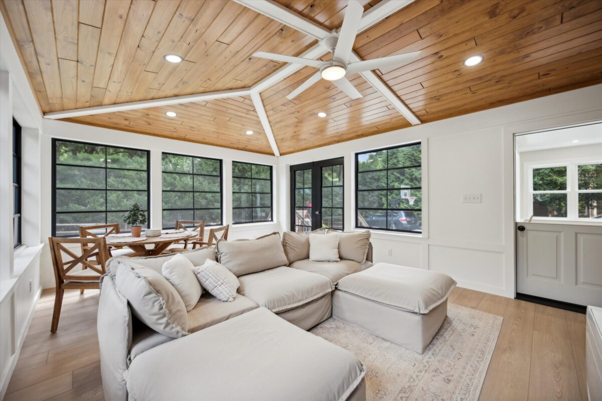 A bright sunroom featuring a comfortable beige sectional sofa and a wooden dining table with chairs. The room has a wooden ceiling, large windows with black frames, and a ceiling fan. Light wood floors and greenery are visible outside.