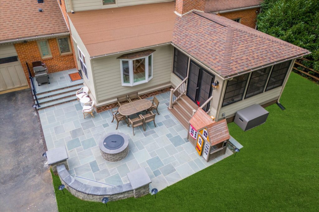 Aerial view of a backyard featuring a stone patio with a round table and chairs, a fire pit, and a small playhouse. The patio connects to a screened porch with steps leading to a well-maintained grassy lawn. The house is brick and siding.