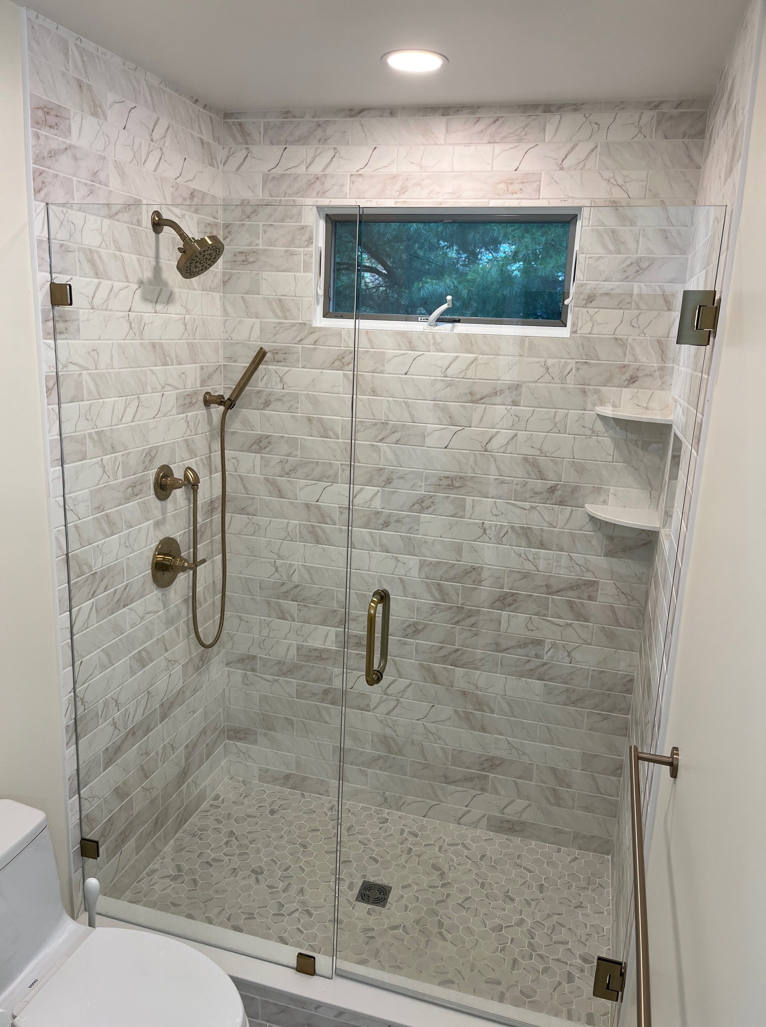 A modern shower with marble-patterned tiles, a glass door, bronze fixtures, and a rainfall showerhead. There is a handheld shower attachment, built-in shelves, and a window providing natural light. The bathroom also features a glimpse of a toilet.