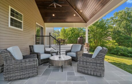 A cozy outdoor patio with four wicker chairs and a round table on a tiled floor. The area is covered by a wooden ceiling with a fan and overlooks a lush green garden. Stairs lead up to a higher deck visible in the background.