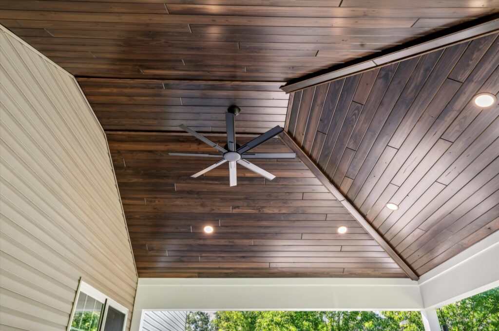 A covered outdoor patio features a vaulted wooden ceiling with recessed lighting and a large, modern ceiling fan. The space is bordered by beige siding and an open view of green trees outside.