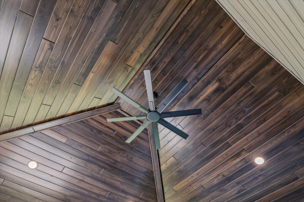 A ceiling with dark wooden panels arranged diagonally, featuring a modern, black, six-blade ceiling fan in the center and two recessed lights on either side.