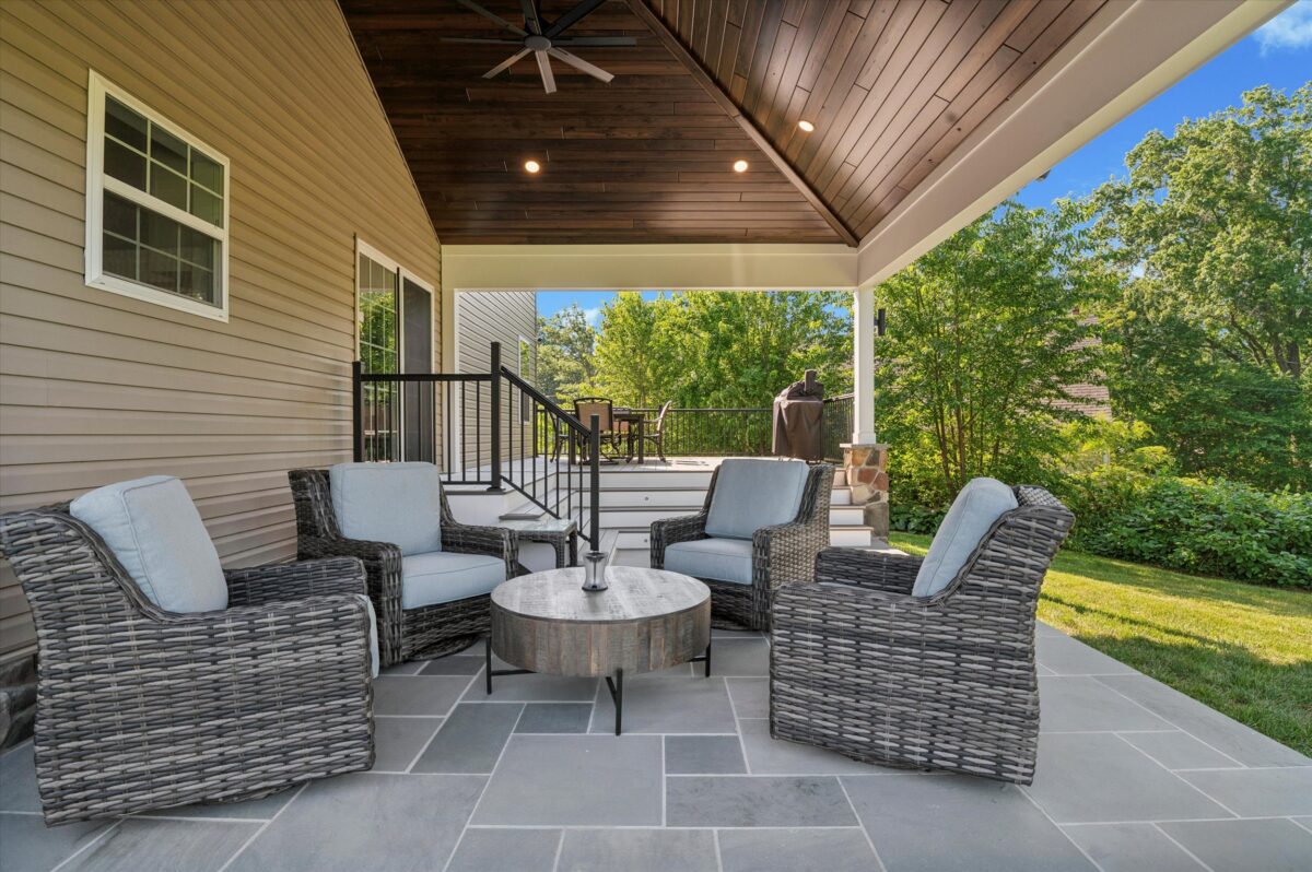 A cozy outdoor patio with four wicker chairs and a round table on a tiled floor. The area is covered by a wooden ceiling with a fan and overlooks a lush green garden. Stairs lead up to a higher deck visible in the background.