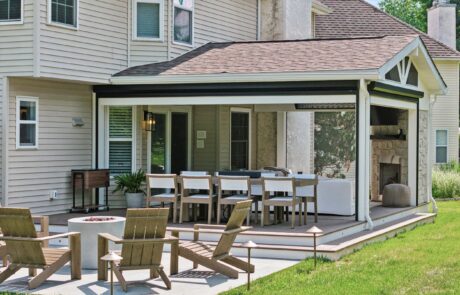 A cozy outdoor patio with a long dining table and chairs under a covered extension. Theres a fireplace and seating area with wooden chairs and a small round fire pit on a concrete area surrounded by grass.