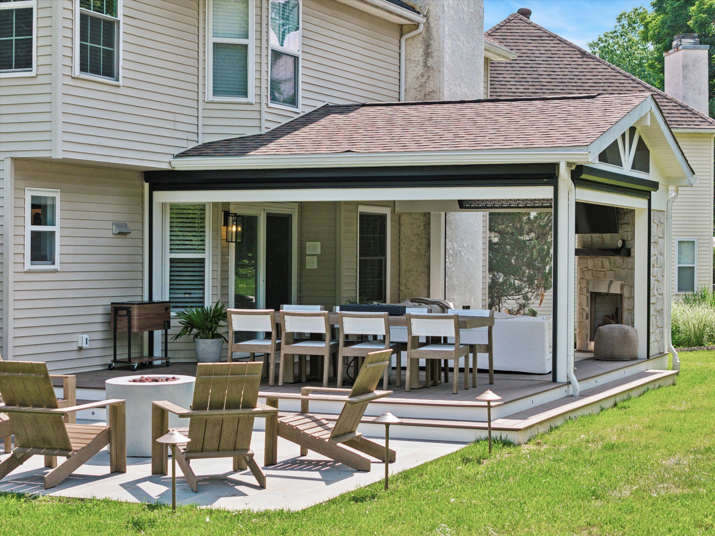 A cozy outdoor patio with a long dining table and chairs under a covered extension. Theres a fireplace and seating area with wooden chairs and a small round fire pit on a concrete area surrounded by grass.