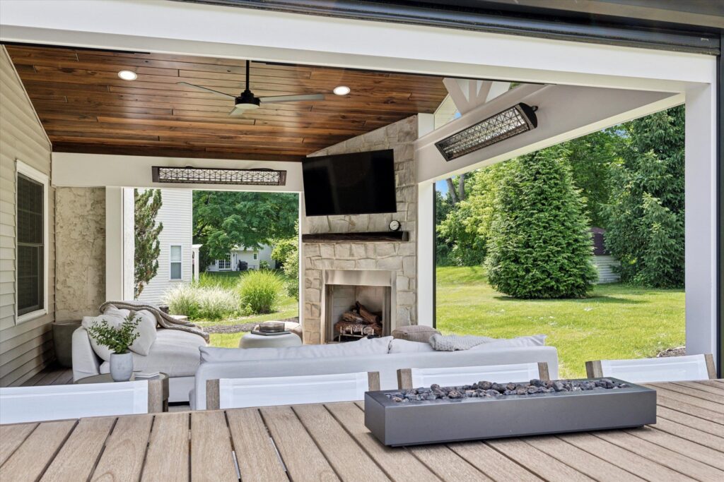 Outdoor patio with white sofas facing a stone fireplace and mounted TV. Ceiling has wood paneling and a fan. Heater lamps are installed above. Large lawn and trees visible in the background. A rectangular fire pit on a wooden deck in the foreground.