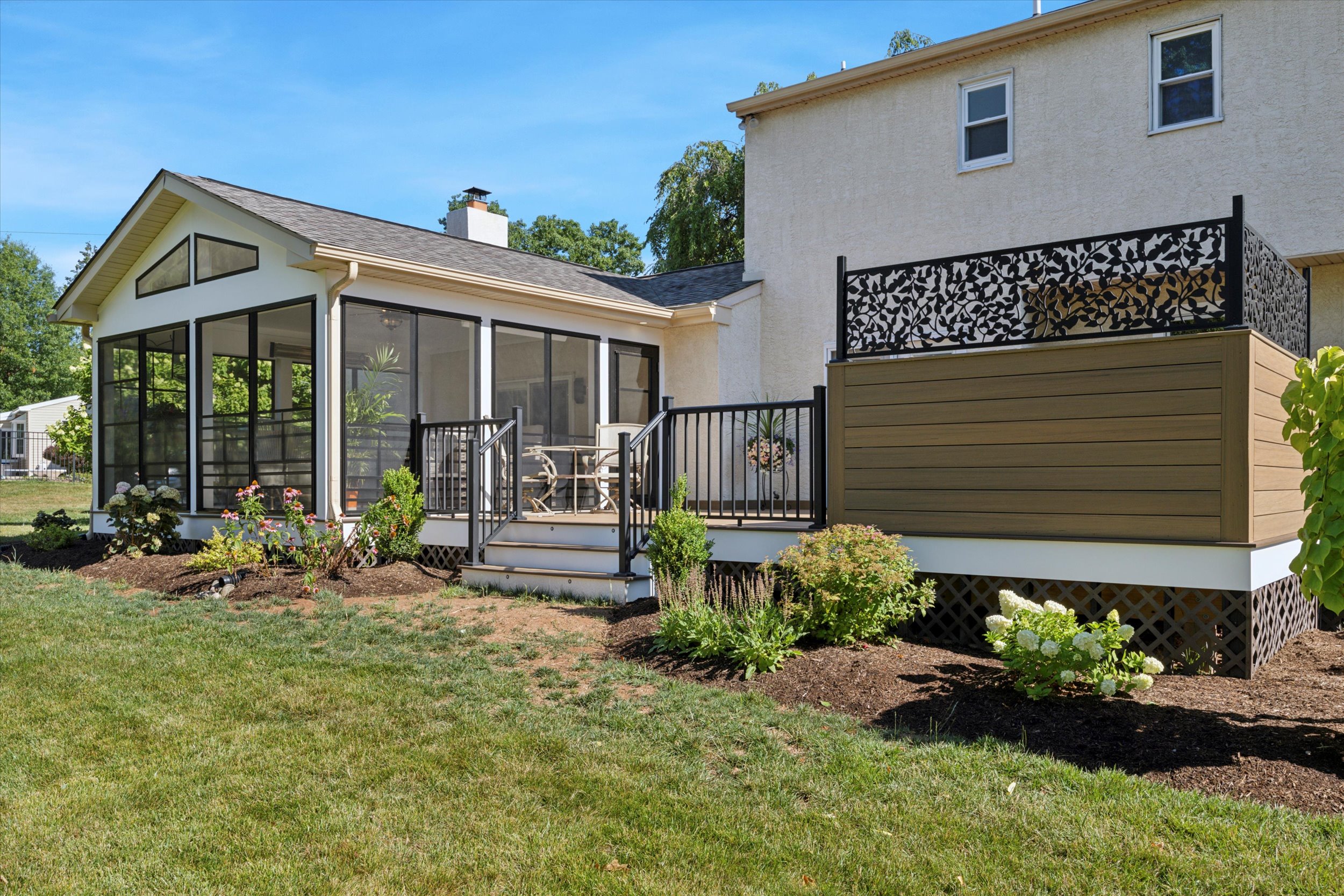 A modern house exterior with a screened porch featuring glass doors and a seating area. The porch has black railings and overlooks a lawn with neatly trimmed bushes. The house is two stories with light-colored siding and a decorative privacy wall.