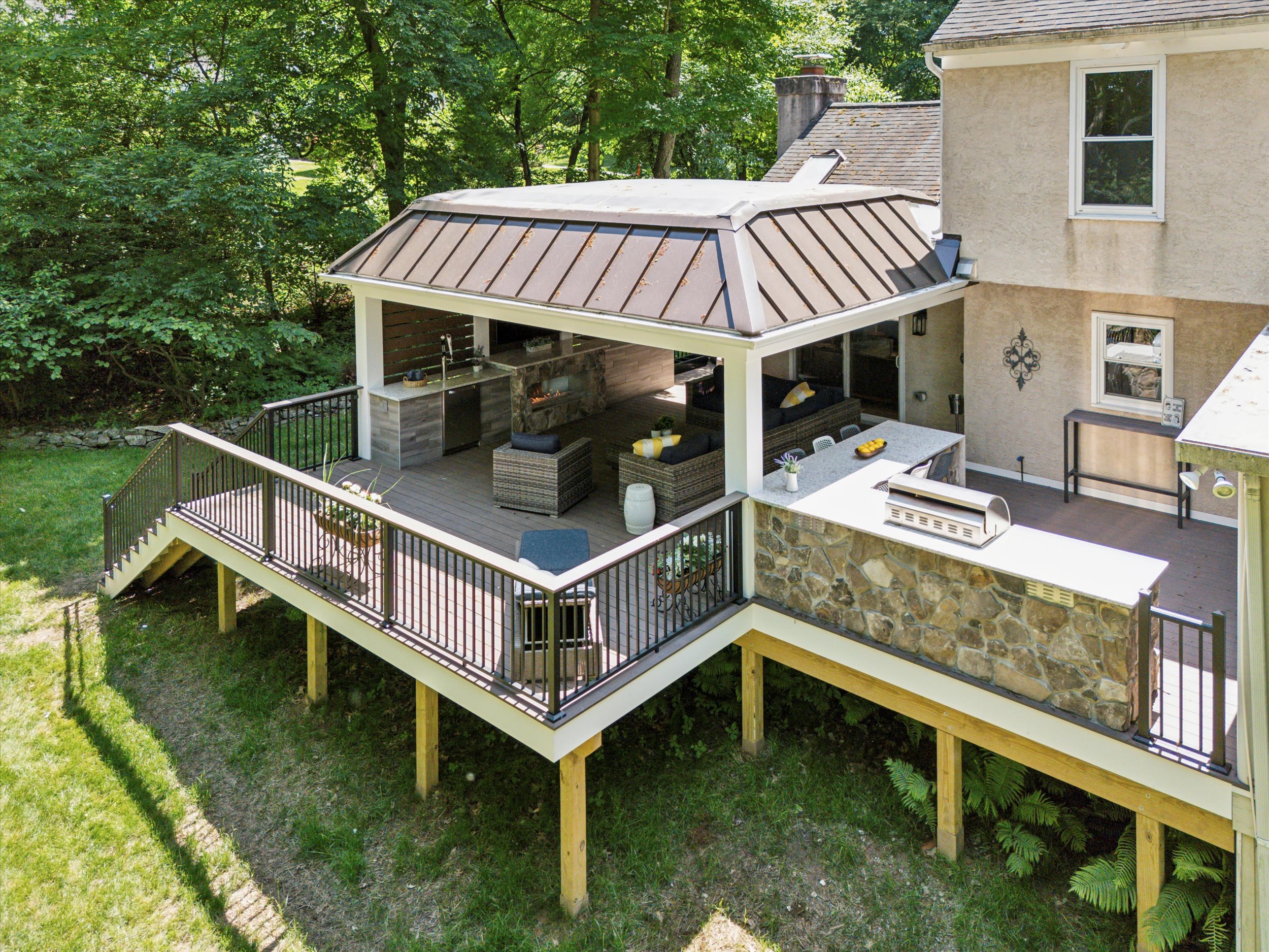 Spacious backyard with a wooden deck featuring a metal roof over an outdoor kitchen and lounge area. Surrounded by lush greenery, it includes cozy seating, a grill, and a bar counter, creating an inviting space for relaxation and entertainment.