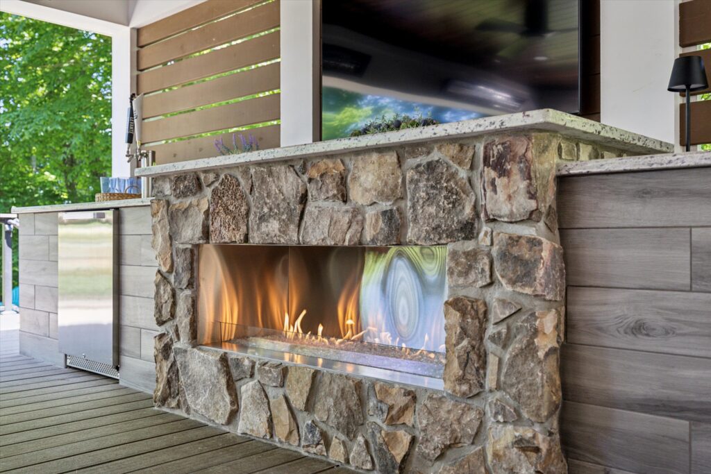 An outdoor fireplace with a rustic stone facade and a sleek, modern linear flame is set on a wooden deck. The area includes a mini fridge and is surrounded by lush green trees, creating a cozy and inviting atmosphere.