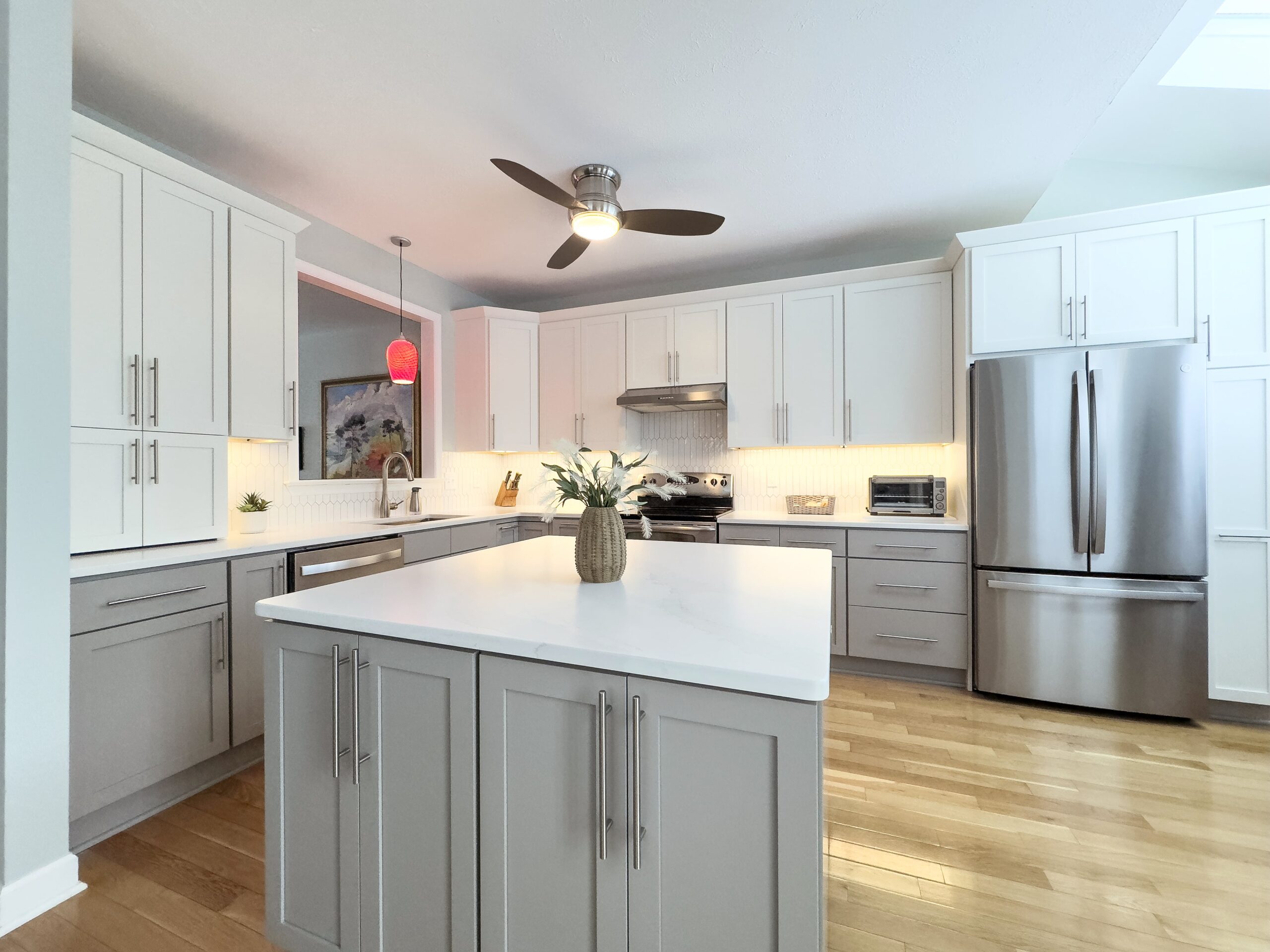 Modern kitchen with gray and white cabinetry, a central island, and stainless steel appliances. A vase with white flowers sits on the island. The kitchen is well-lit with a ceiling fan above and a red pendant light by the counter. Wood flooring throughout.