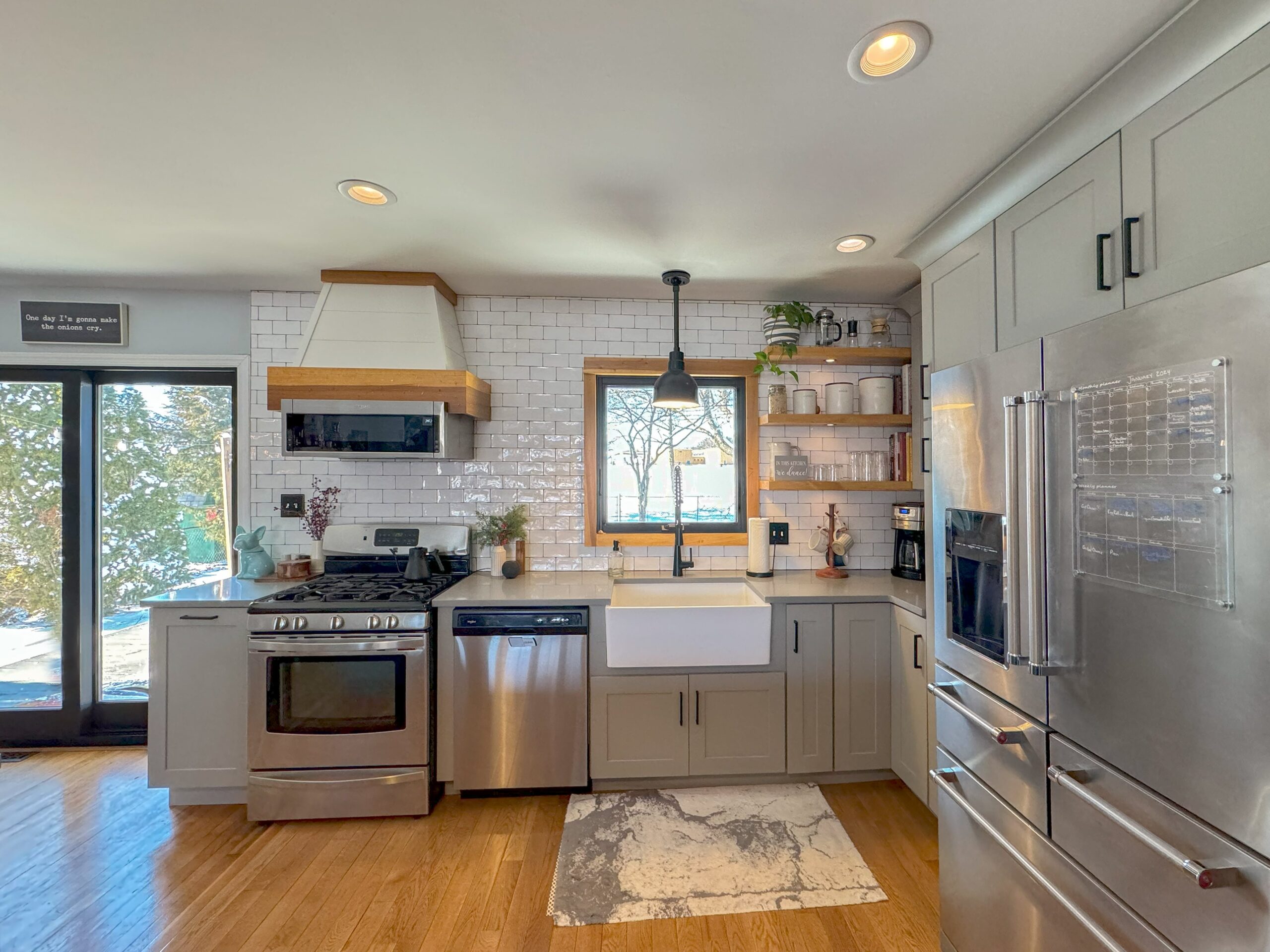 A modern kitchen with stainless steel appliances, white subway tile backsplash, and light gray cabinets. A large farmhouse sink is under a window. Open shelves display dishes, and a rug lies on the wooden floor. Glass doors reveal a snowy scene outside.