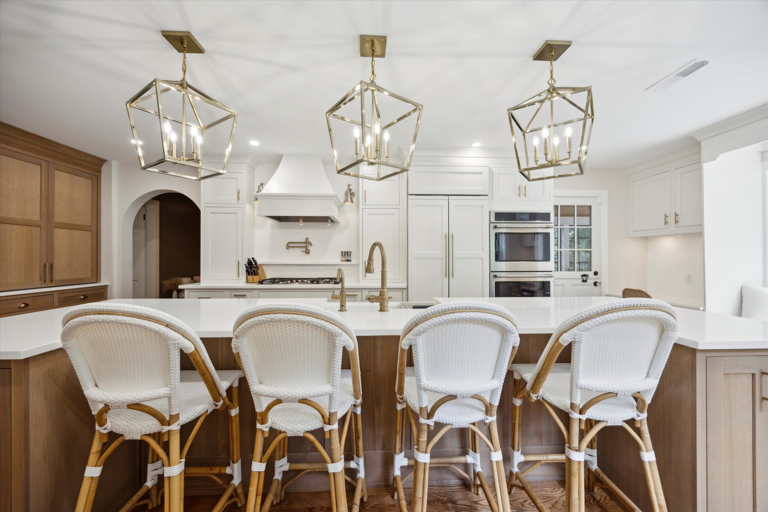 A modern kitchen with wooden flooring features white cabinets and a central island with four wicker bar stools. Three geometric pendant lights hang above the island, with stainless steel appliances and an arched doorway in the background.