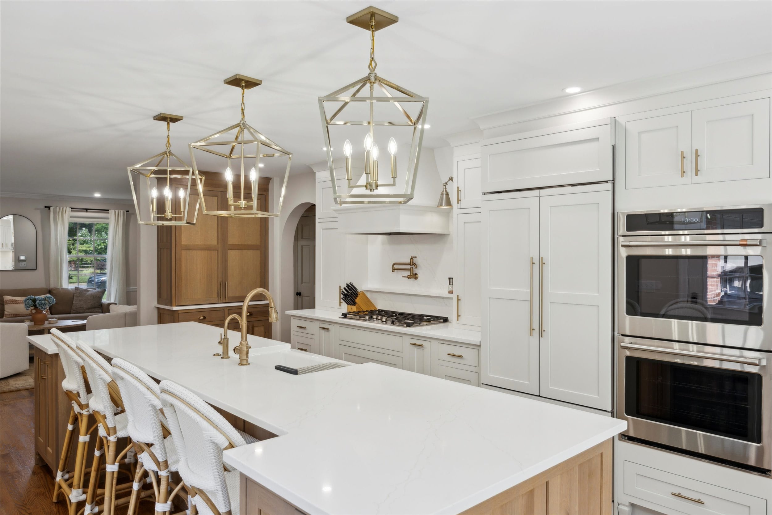 Modern kitchen with white cabinets and a large island featuring wicker bar stools. Geometric gold pendant lights hang above, and stainless steel double ovens are built into the wall. An adjacent living area is visible in the background.