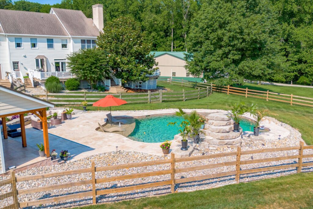 A backyard view featuring a small in-ground pool with a rock waterfall and lounge chair under an orange umbrella. The pool area is surrounded by a wooden fence, trees, and a two-story white house. Theres also a patio with potted plants.