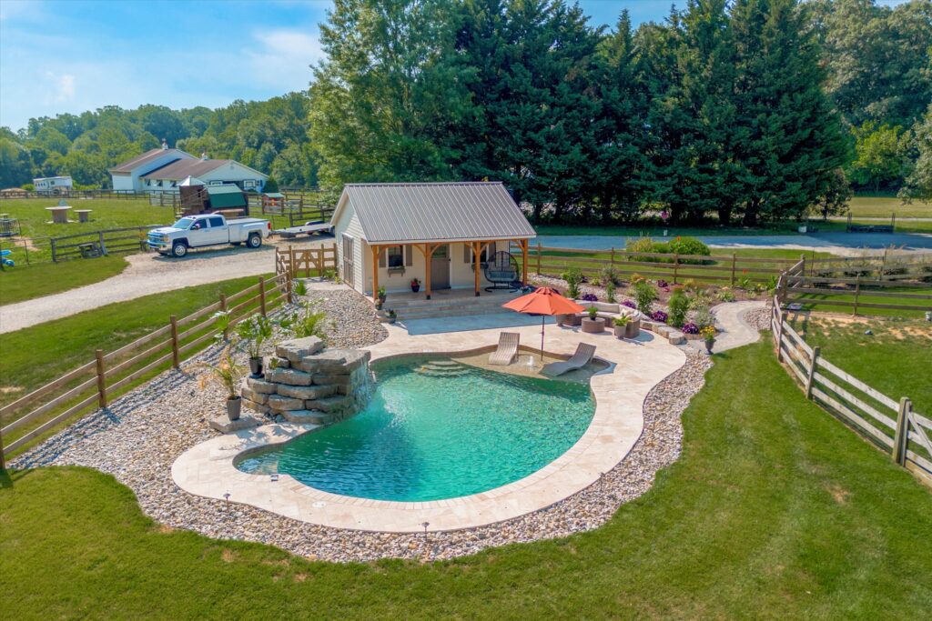 A tranquil backyard scene with a kidney-shaped pool, surrounded by stone paving. A small waterfall feature flows into the pool. Nearby, an orange umbrella shades two lounge chairs. A cozy pool house sits in the background, with lush greenery around.