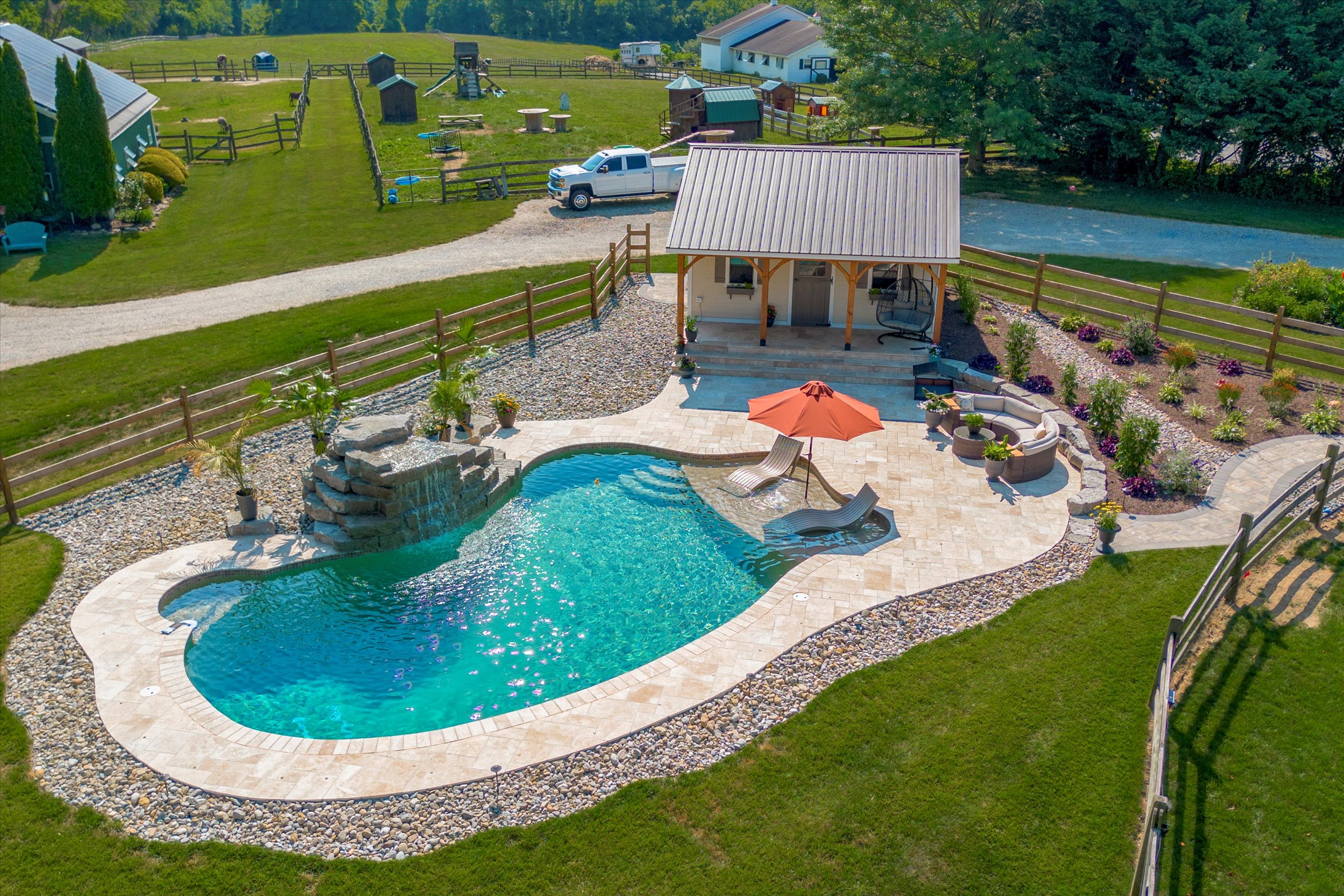 Aerial view of a curvy swimming pool with a rock waterfall feature and a surrounding patio. Theres a small pool house, lounge chairs with an orange umbrella, a stone fire pit, and landscaped gardens, all enclosed by a wooden fence.