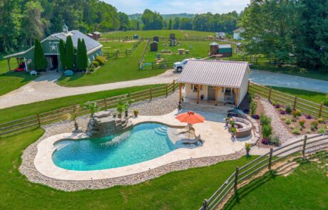 A scenic rural property with a kidney-shaped pool, surrounded by a stone deck. An umbrella and lounge chairs sit poolside. A small cabana and lush green lawn extend outwards, with trees and a barn visible in the background.