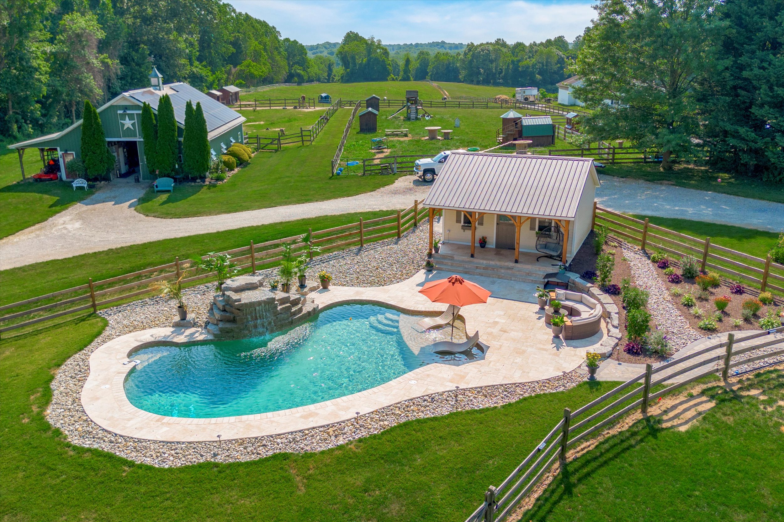 A scenic rural property with a kidney-shaped pool, surrounded by a stone deck. An umbrella and lounge chairs sit poolside. A small cabana and lush green lawn extend outwards, with trees and a barn visible in the background.